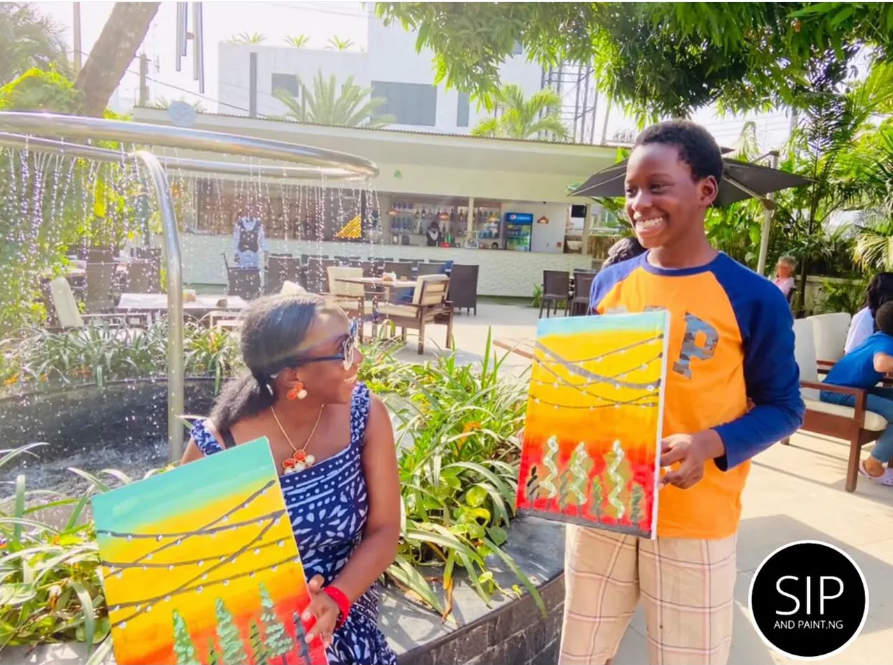 A mother and son at Cilantro Abuja Maitama smiling with their matching sunset paintings beside the garden fountain