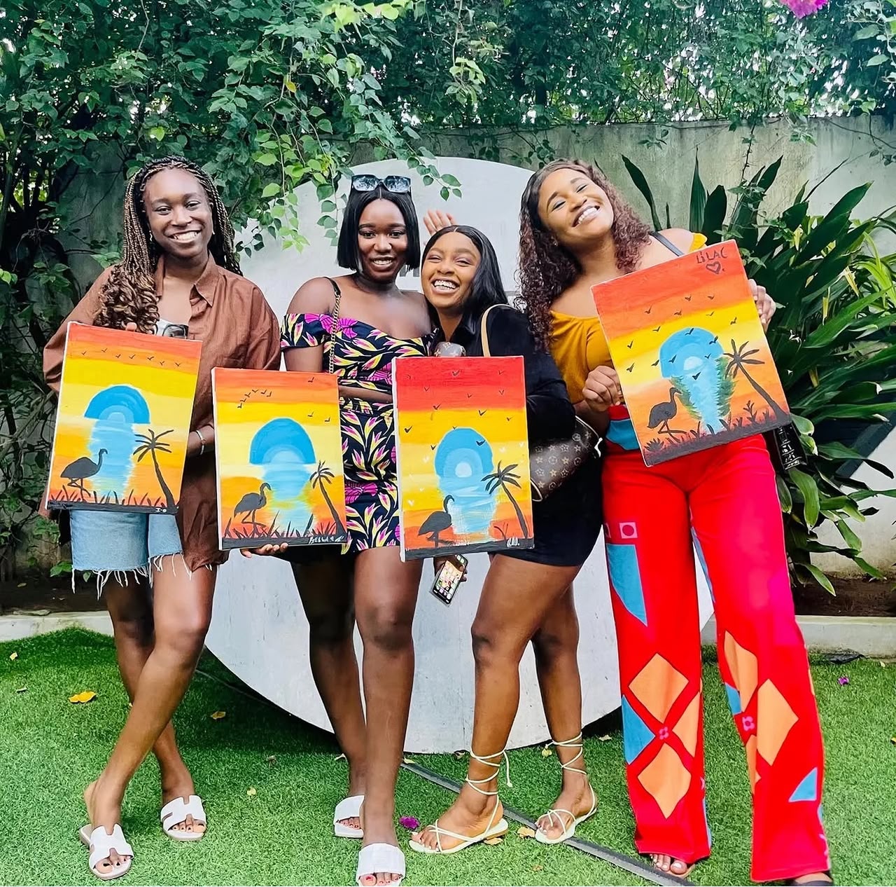 Four friends in an Abuja garden holding matching sunset paintings with palm trees and herons after a Sip and Paint session
