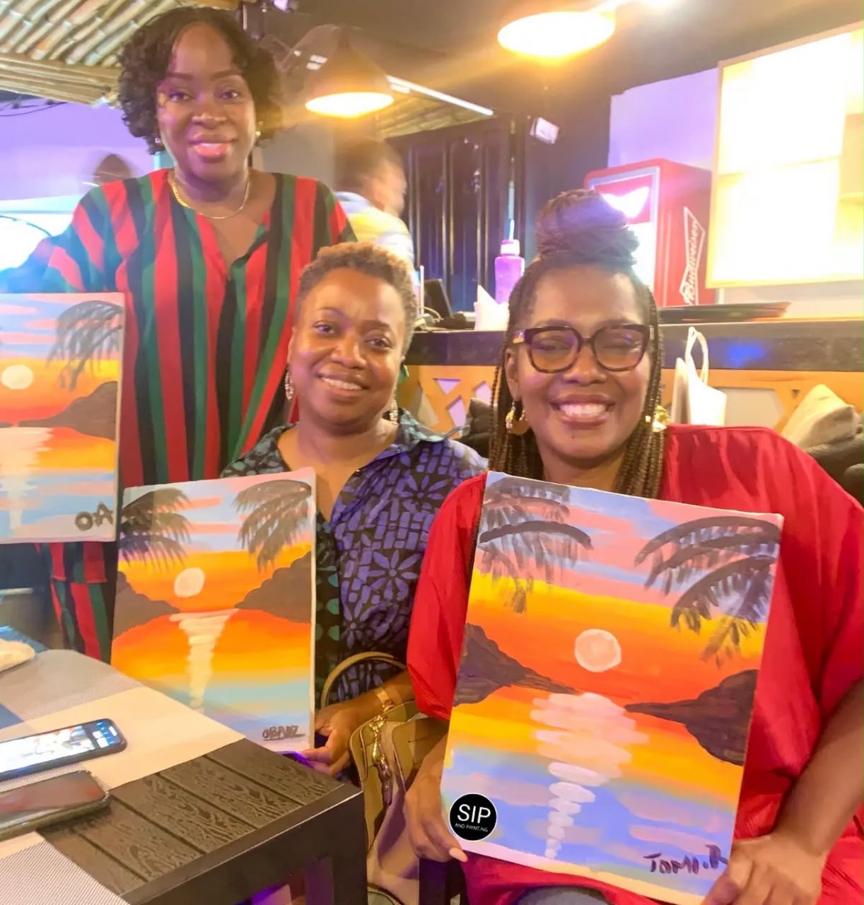 Three women at Blackstone Gwarinpa holding matching palm tree sunset paintings after a Sip and Paint session