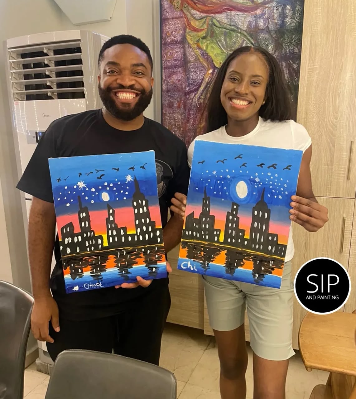 A couple in Abuja holding matching city-skyline canvas paintings after a Sip and Paint session