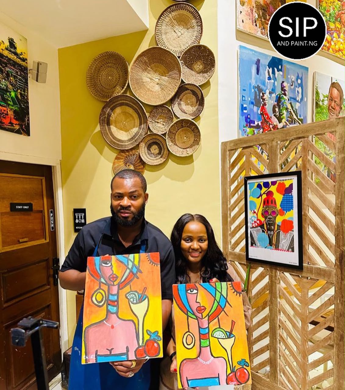 A couple at Cilantro Abuja Maitama holding vibrant abstract portrait paintings beside a wall of woven baskets