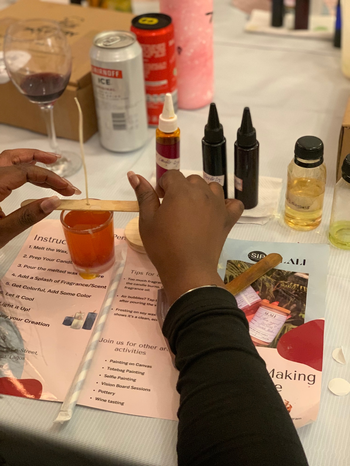 Hands at a Sip and Paint candle making session, wick balanced on a wooden stick over a glass vessel of amber wax beside dye bottles, a wine glass and instructions